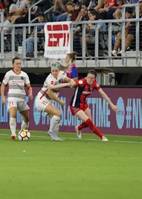A competitive soccer match is underway with players vying for the ball near the sideline. The players wear different uniforms, indicating they are from opposing teams. The audience is visible in the stadium seating above, and there are visible advertisements and hashtags on the LED boards along the field.
