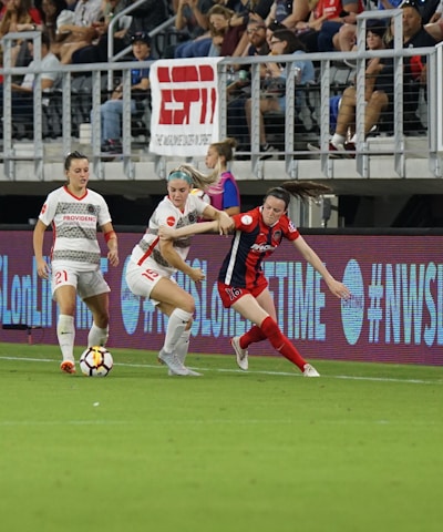 A competitive soccer match is underway with players vying for the ball near the sideline. The players wear different uniforms, indicating they are from opposing teams. The audience is visible in the stadium seating above, and there are visible advertisements and hashtags on the LED boards along the field.