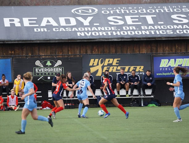 Players engage in a competitive soccer game on a field while others are seated on the bench watching attentively. The action is taking place in front of a wooden structure displaying multiple advertisements, including orthodontics, lacrosse, and field hockey.