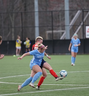 Players engaged in a competitive soccer match on a green field. One player in a blue uniform is actively kicking the ball while another player in a red and black uniform attempts to block. Two additional players in blue uniforms and another player in a red uniform are closely observing the action. The background shows others standing near the sideline.