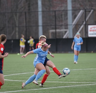 Players engaged in a competitive soccer match on a green field. One player in a blue uniform is actively kicking the ball while another player in a red and black uniform attempts to block. Two additional players in blue uniforms and another player in a red uniform are closely observing the action. The background shows others standing near the sideline.
