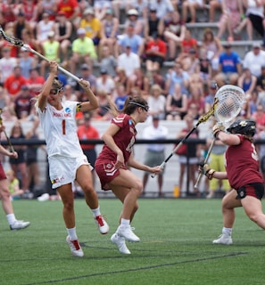 Athletes engaged in a competitive lacrosse match on a grassy field, with one player in white attempting to score against a goalie in burgundy gear. The scene is lively, with a full audience in the background, dressed casually and attentively watching the game.