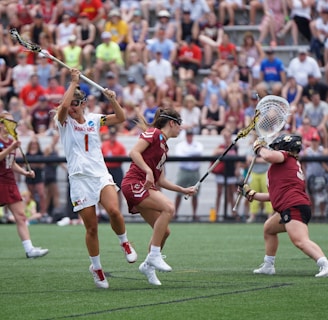 Athletes engaged in a competitive lacrosse match on a grassy field, with one player in white attempting to score against a goalie in burgundy gear. The scene is lively, with a full audience in the background, dressed casually and attentively watching the game.