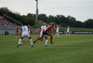 A competitive women's soccer match taking place on a grassy field. Players in red and black are attempting to gain possession of the ball, while players in white are defending. The scene conveys action and movement, with players focused on the game. Spectators are seated in the background, and there are advertisements along the sidelines.