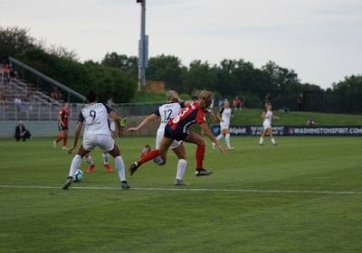 A competitive women's soccer match taking place on a grassy field. Players in red and black are attempting to gain possession of the ball, while players in white are defending. The scene conveys action and movement, with players focused on the game. Spectators are seated in the background, and there are advertisements along the sidelines.