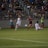 A competitive soccer match is taking place with players in action on the field. Two players in white uniforms are being challenged by an opponent in a red and black uniform. The crowd in the background is seated in bleachers, watching the intense play. The field is well-lit, and a ball is in motion near the players.