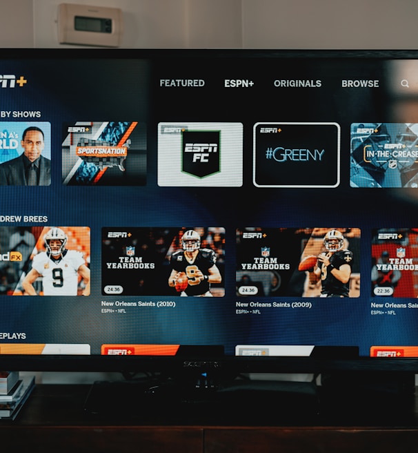 A television screen displaying the ESPN+ streaming interface with various sports-related shows and categories. The screen includes sections such as 'Browse by Shows' and 'Best of Drew Brees' with thumbnails of sports programs featuring athletes in action and analysts.